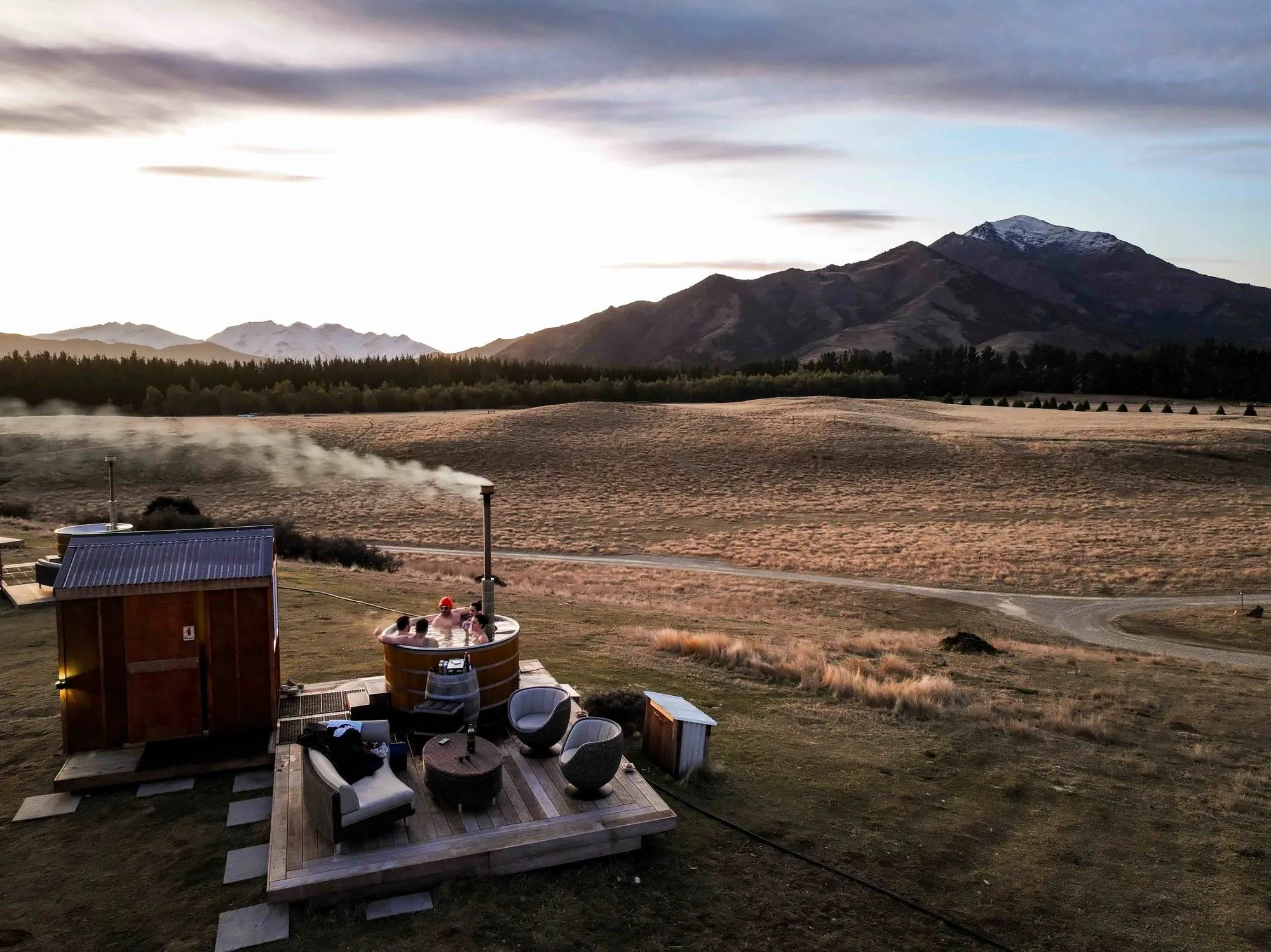 Friends soaking in a steaming hot tub on a timber deck at sunset, with tussock farmland and snow-capped peaks behind